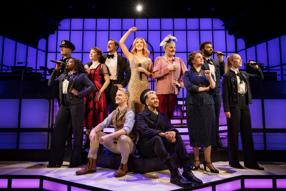A diverse group of performers on a lit theater stage, some standing and two kneeling in front, with a woman in a gold sequin dress center holding a microphone amid purple stage lighting.