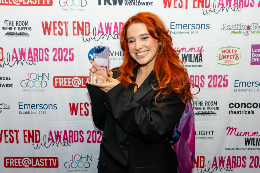 Smiling woman with red hair holding a glass award at the West End Awards 2025 backdrop.