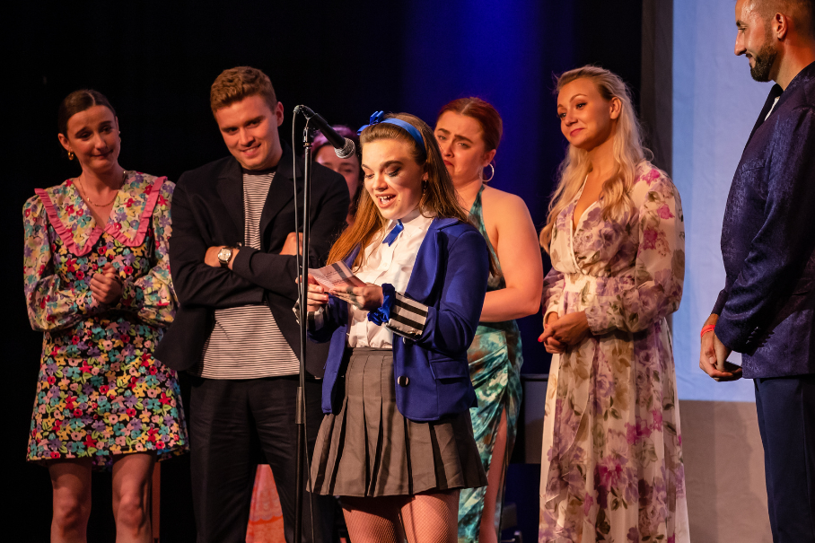 Young woman in a blue jacket speaks into a mic on stage, flanked by friends watching and smiling as others stand in the spotlight.