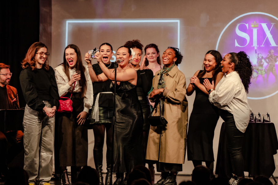 Group of diverse women on a stage celebrating, some posing for a selfie while others cheer, with a circular logo visible in the background.