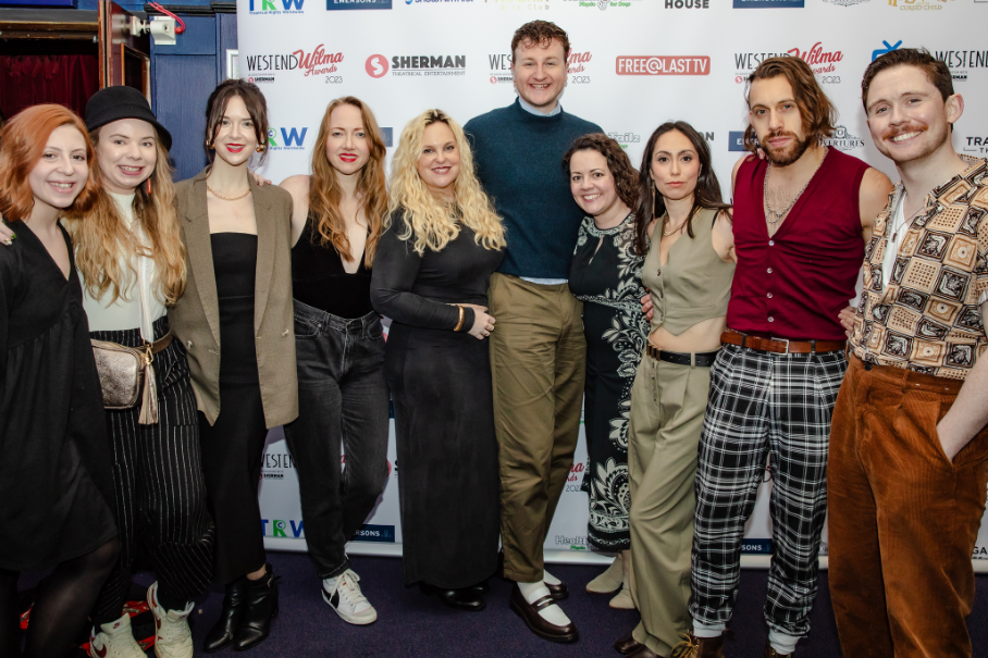 Group photo of about a dozen people posing on a blue carpet at a media event, smiling in varied outfits with a logo-covered backdrop behind them.