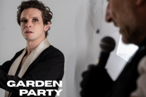Person in black with a light scarf looks toward the camera as another person speaks into a microphone (blurred) in the foreground; 'GARDEN PARTY' text overlaid.