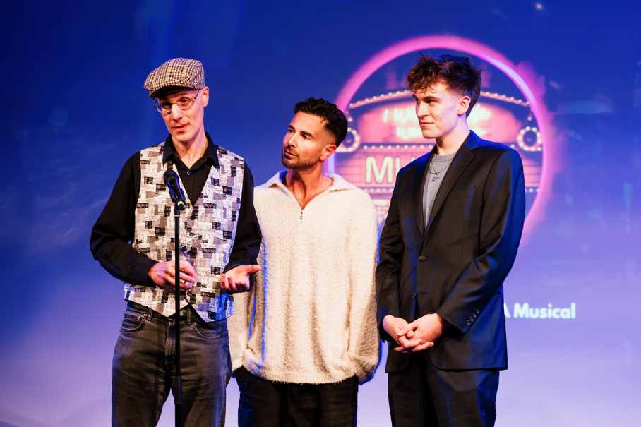 Three men stand on a stage, the left speaker wearing a flat cap and patterned vest while speaking into a microphone.
