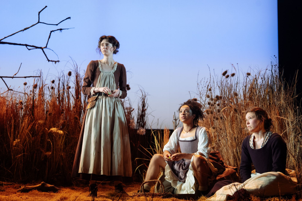 Three women in period costumes on a stage set as a field, with one standing and two seated, looking upward or thoughtful.