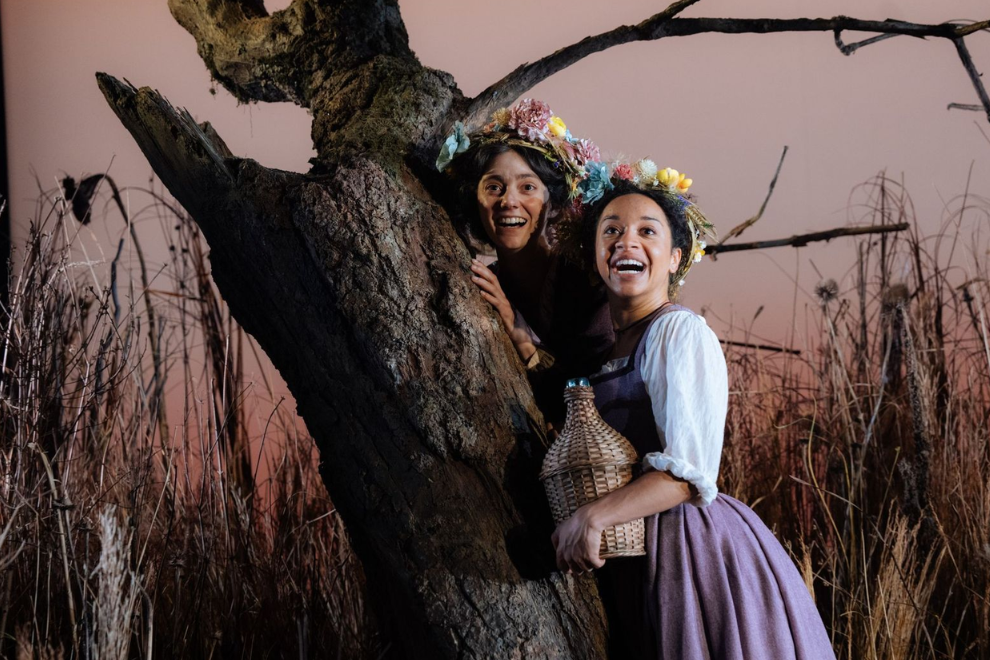 Two women wearing flower crowns smile and lean against a large tree in a dry field at sunset; one holds a wicker-wrapped bottle.