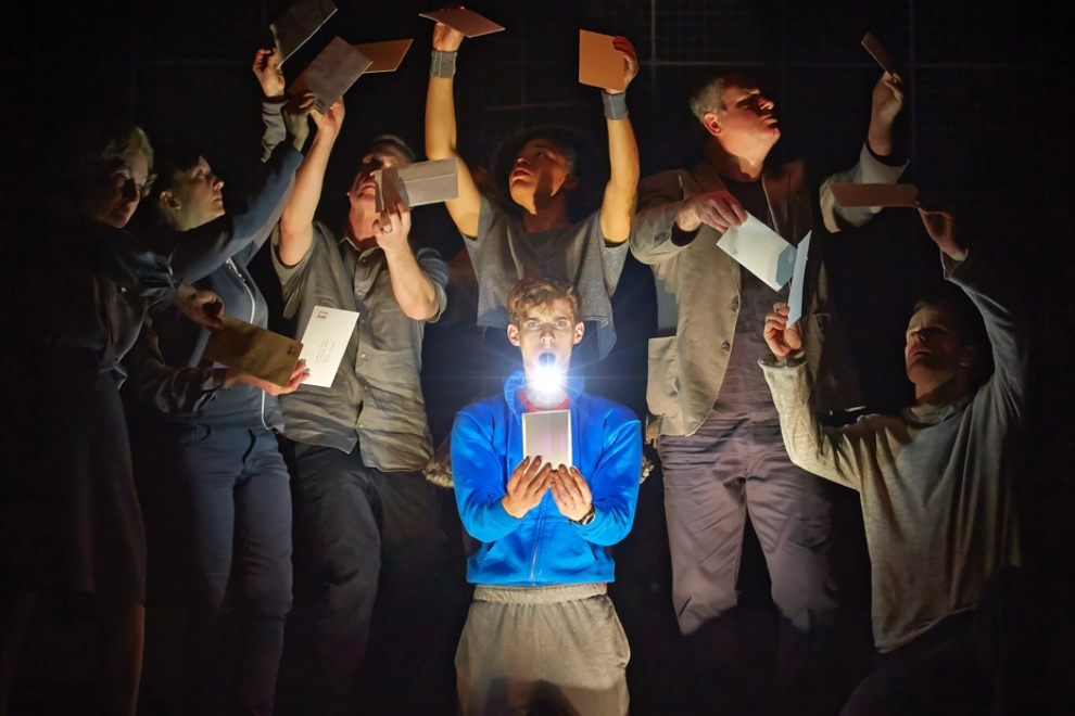Young man in a blue jacket holds a glowing box, center of a group of people in a dark room, envelopes flying around him as lights shine from the box.