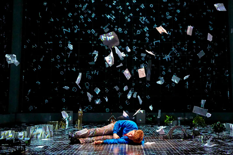A person in a blue shirt lies on a dimly lit stage as papers with letters float around them.