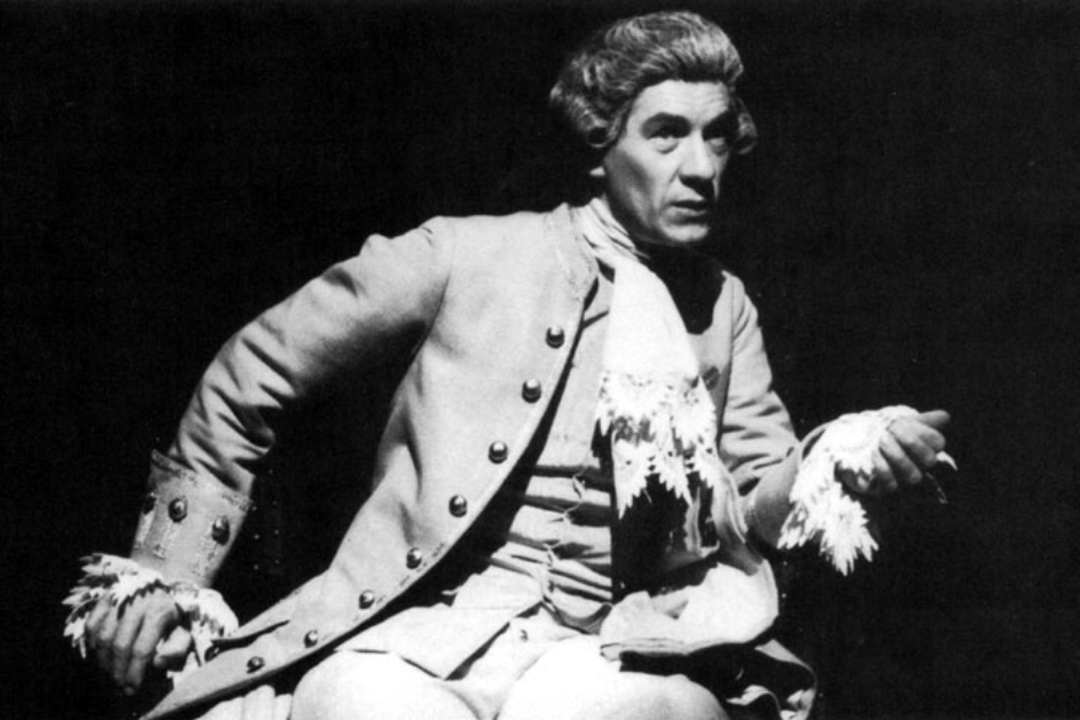 Actor in an 18th-century costume performing on stage, gesturing with fringed cuffs in a black-and-white scene