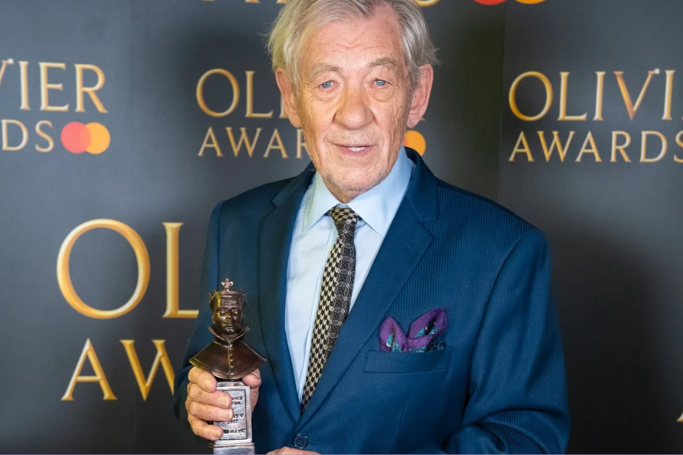 Older man in a blue suit holding a small statue trophy at an awards ceremony, with 'OLIVI AWARDS' backdrop in gold letters.