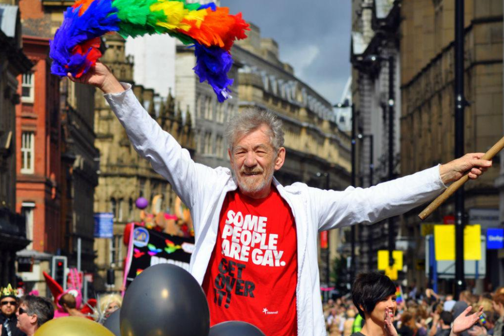 Older man with gray hair wearing a red shirt that reads 'Some people are gay. Get over it!' raises a rainbow feather garland above his head during a Pride parade on a city street, with cheering crowd and historic buildings in the background.