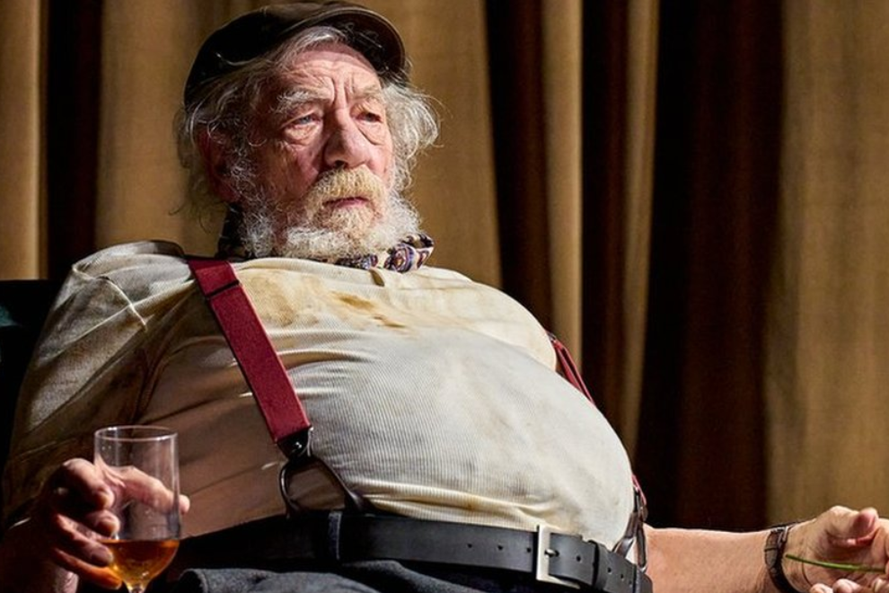 An elderly man with a white beard wearing a cream shirt and red suspenders sits holding a whiskey glass on stage.