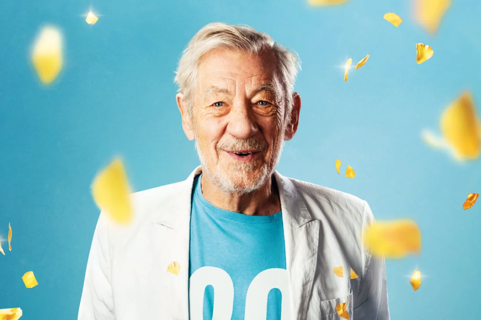 Smiling elderly man in a white blazer and turquoise shirt amid yellow confetti against a blue background.