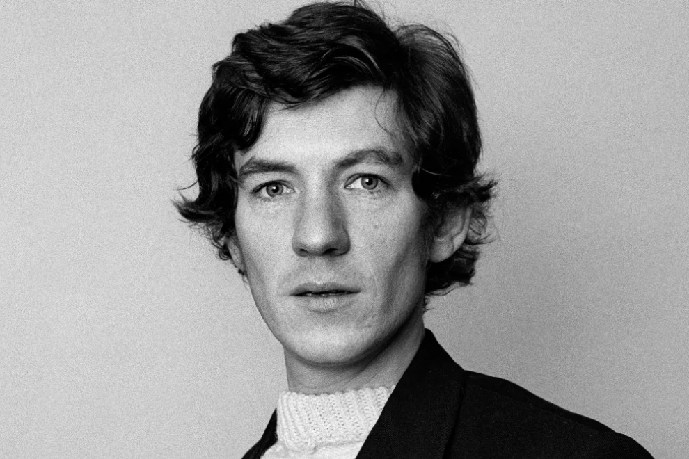 Black-and-white head-and-shoulders portrait of a man with wavy dark hair, wearing a blazer and white knit turtleneck, looking at the camera.