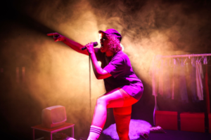 Vibrant concert scene: a vocalist in purple outfit sings into a microphone, one knee raised, bathed in orange stage lights.