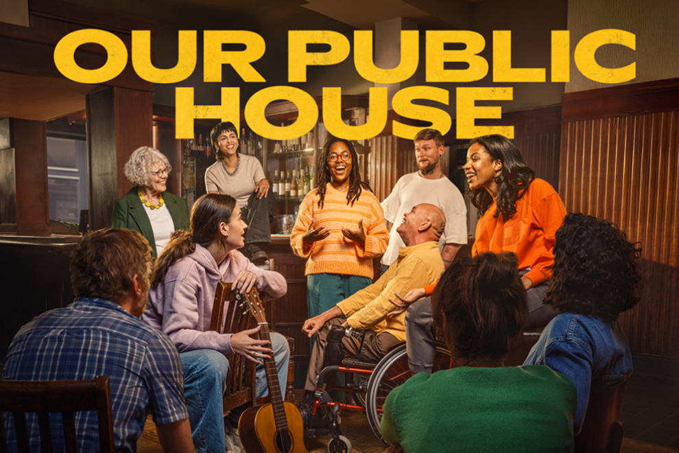 Diverse group of people in a circle, including a person in a wheelchair, talking in a cozy room with a large yellow 'OUR PUBLIC HOUSE' banner overhead.