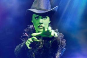 Green-skinned witch in a tall hat reaches toward the viewer under blue lighting, casting a dramatic spell-like pose.
