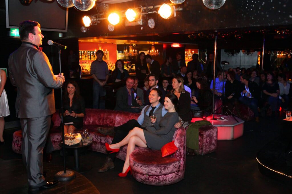 A man in a gray suit sings into a microphone on a small stage while a crowded lounge watches from velvet sofas and a bar backs the scene.