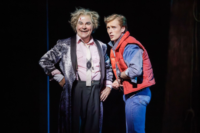 Two male actors on a dark stage; one in a shiny patterned robe and pink shirt, the other in a red vest and blue jeans.