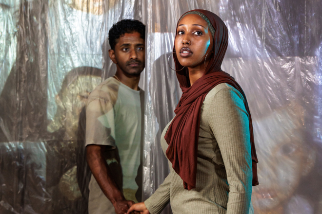 Two people stand in front of a translucent plastic wall; a woman wearing a brown headscarf and beige outfit looks toward the camera while a man in a light T‑shirt stands behind her, both with serious expressions.