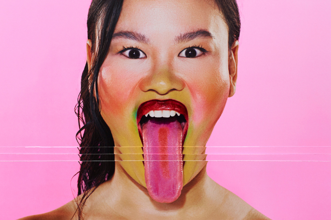 Close-up portrait of a person with rainbow makeup, sticking out a bright pink tongue against a pink background.