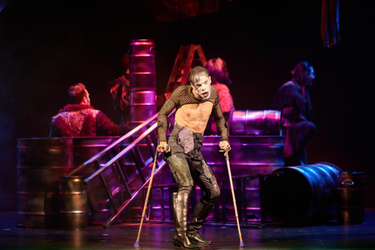 Performer with white skeleton makeup on stage, leaning on crutches during an industrial-themed theatrical scene with barrels and ladders in the background.