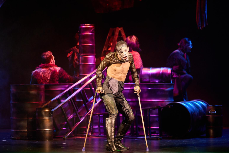 Performer with white skeleton makeup on stage, leaning on crutches during an industrial-themed theatrical scene with barrels and ladders in the background.