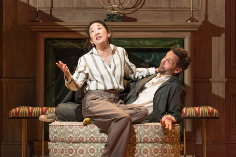 Woman in a striped blouse gestures dramatically as she sits on a patterned ottoman on stage, a man in a blazer listening beside her.