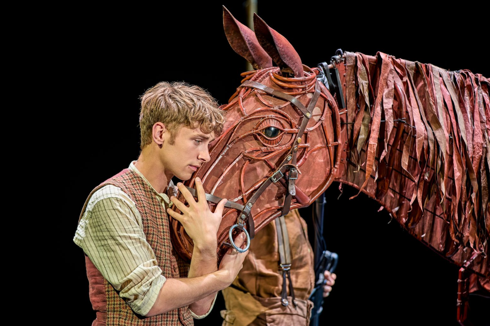 Young man in period costume gently touching a leather horse prop on a stage set against a dark background, suggesting a theater performance.