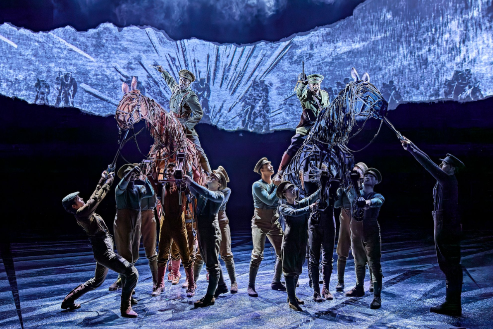 Theatrical scene with multiple performers guiding two large horse puppets on a dark blue-lit stage.