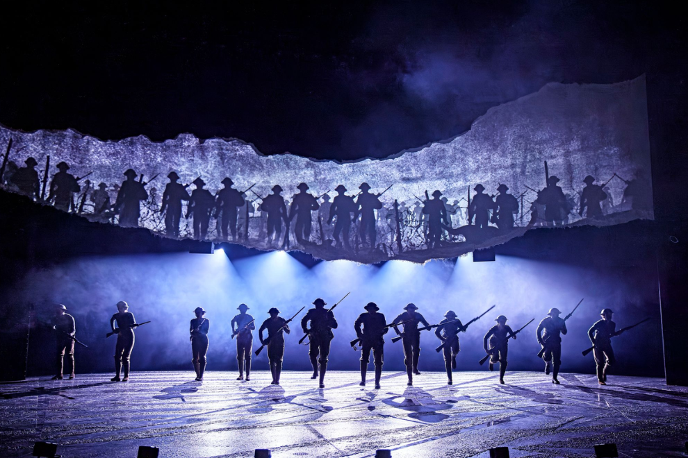 Silhouetted soldiers marching on a dimly lit stage with a large troops-projection backdrop and blue spotlights. Stage scene of military figures in formation against a mountainous silhouette, bathed in blue light and fog. A line of performers in uniform carry rifles as they march, with a shadowy army image projected above and dramatic blue lighting.