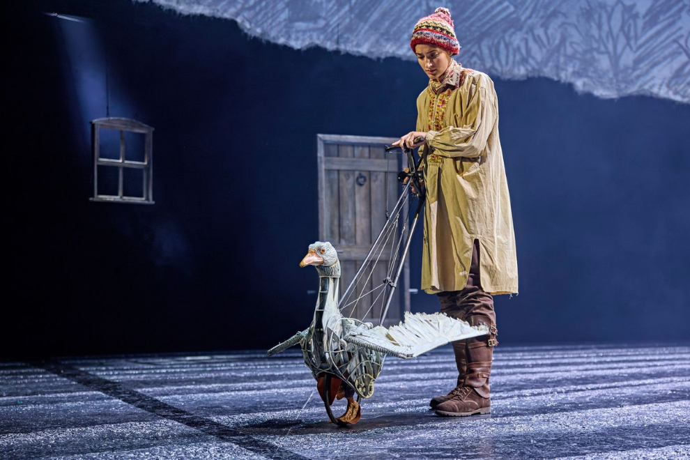 Puppeteer in a beige coat and knit hat controls a wheeled, string-operated turkey puppet on a dark stage.