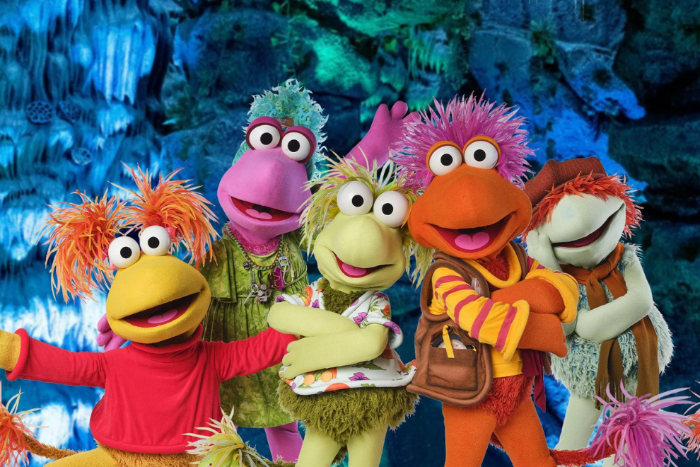 Six colorful furry puppets pose together for a group photo against a blue cavern backdrop, smiling widely with arms around each other and bright outfits.