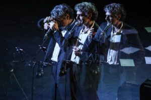 Three male singers in suits perform on a dimly lit stage, multiple-mic setup and motion-blur overlays.