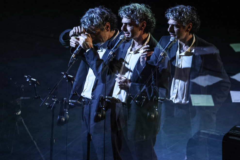 Three male singers in suits perform on a dimly lit stage, multiple-mic setup and motion-blur overlays.