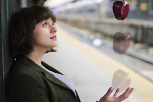Woman at an outdoor train platform gazes upward at floating red apples in the air beside her, creating a surreal moment.