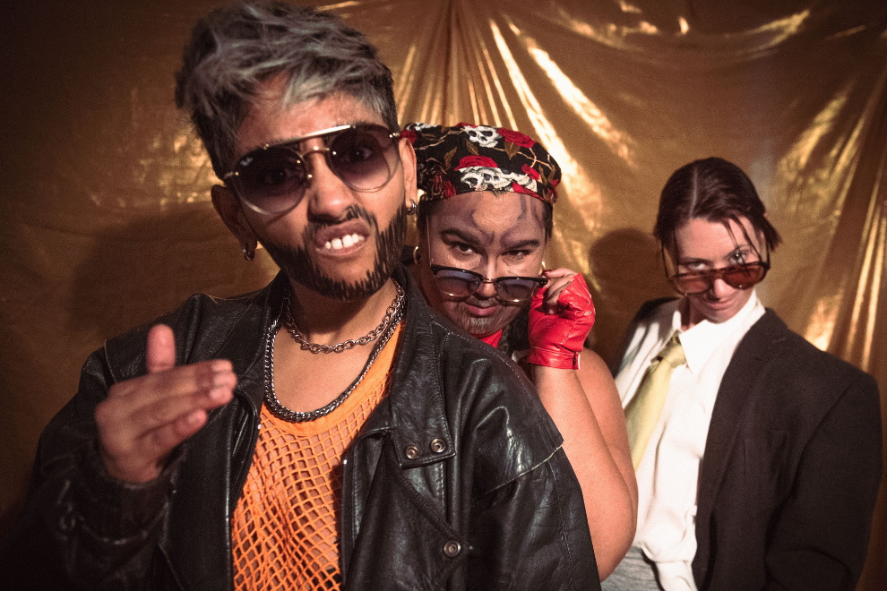 Three friends posing in a retro-fun vibe: man in leather jacket and mesh shirt front, woman with skull bandana and red gloves behind him, another person in a suit and sunglasses to the side.