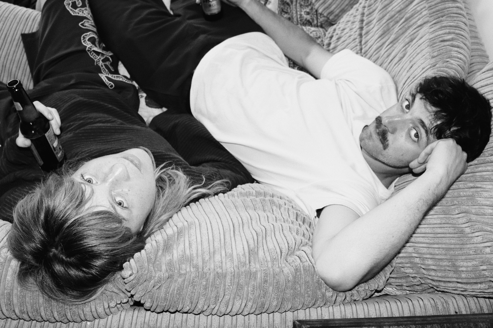 Two people lounging on a striped couch, each holding a beer bottle, looking at the camera.