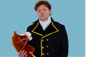 Man in a black double-breasted coat with gold trim holds a brown plush horse against a solid blue background.