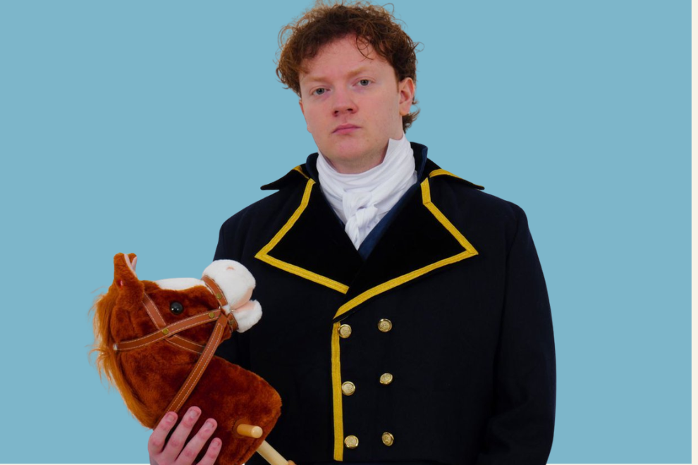 Man in a black double-breasted coat with gold trim holds a brown plush horse against a solid blue background.