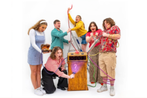 Six people stand around a wooden box with a digital timer and colorful wires, one person pressing a purple-glowing lever as others react with excitement or surprise.