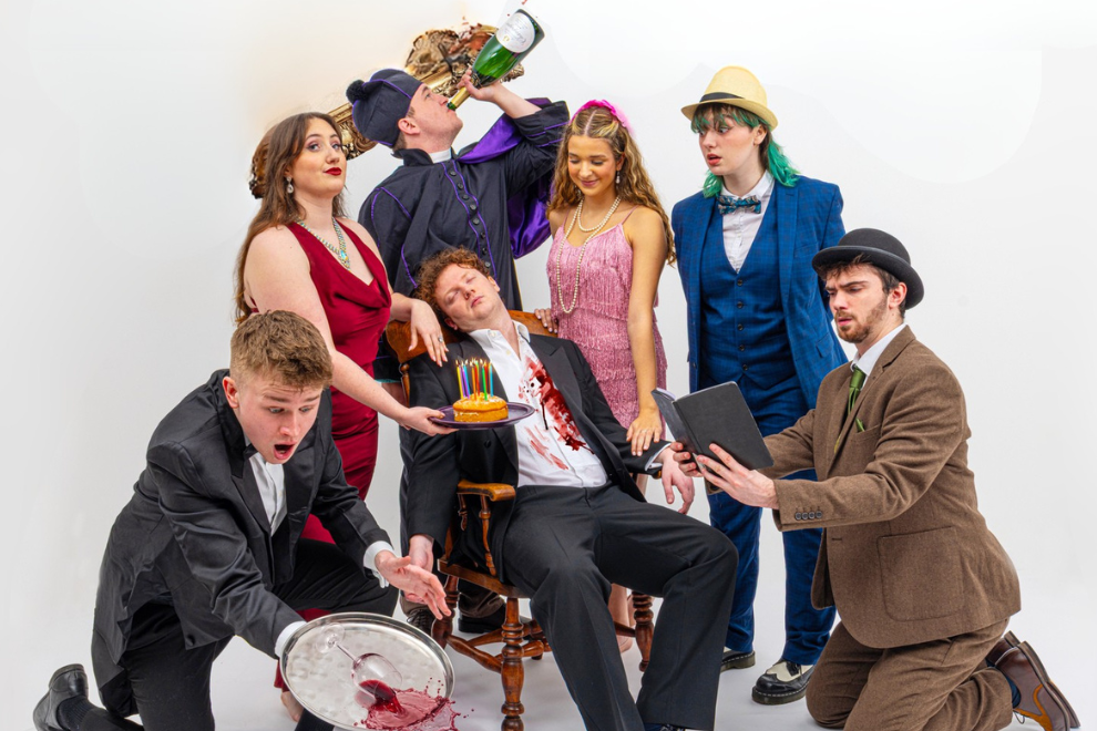 A group of friends in colorful 1920s and formal costumes posing at a party; one man sits slumped with a faux blood-streaked shirt while others peek and toast with cake and drinks.
